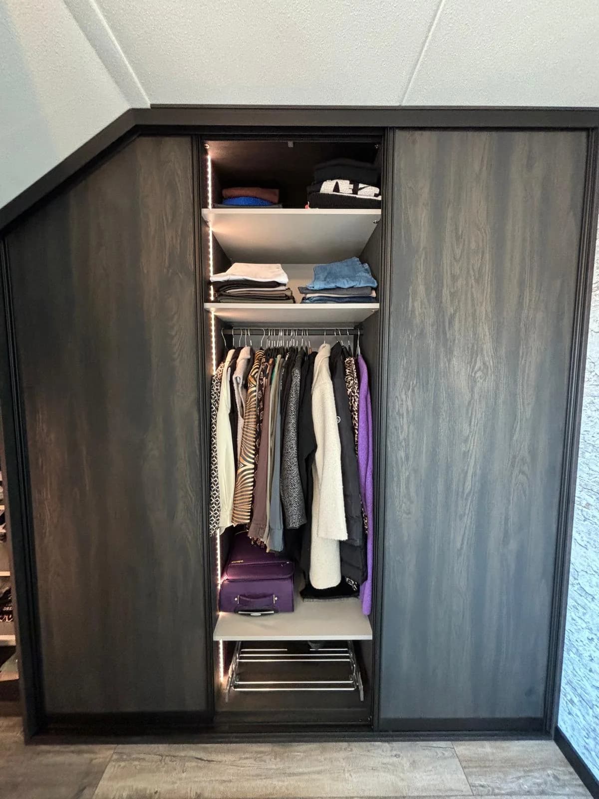 Kledingkast in donker eiken onder een schuin dak, open met kleding
